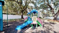 Located at 2220 Gloria Cir in Pensacola, Florida, this single-level outdoor space is centered around a vibrant, child-friendly playground area. The space is defined by its lush, mature oak trees that form a natural canopy overhead, providing generous shade across the grounds. The playground features multiple colorful play structures, including two distinct multi-level units equipped with slides, ramps, and covered platforms. One structure showcases a blue slide and pink accents, while a second unit offers a teal slide alongside a sheltered tower, giving children varied options for active play. The ground beneath the equipment is covered with a layer of dark mulch, providing a safe and well-maintained surface. The entire area is enclosed by a white privacy fence, creating a secure and defined boundary that enhances both safety and the overall aesthetic of the space. Surrounding greenery, including ornamental grasses and the established trees, adds a natural, welcoming atmosphere. The combination of durable, colorful equipment and a thoughtfully maintained natural setting makes this outdoor space an engaging and safe environment for children.