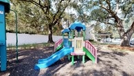Located at 2220 Gloria Cir in Pensacola, Florida, this single-level outdoor space is centered around a vibrant, child-friendly playground area. The space is defined by its lush, mature oak trees that form a natural canopy overhead, providing generous shade across the grounds. The playground features multiple colorful play structures, including two distinct multi-level units equipped with slides, ramps, and covered platforms. One structure showcases a blue slide and pink accents, while a second unit offers a teal slide alongside a sheltered tower, giving children varied options for active play. The ground beneath the equipment is covered with a layer of dark mulch, providing a safe and well-maintained surface. The entire area is enclosed by a white privacy fence, creating a secure and defined boundary that enhances both safety and the overall aesthetic of the space. Surrounding greenery, including ornamental grasses and the established trees, adds a natural, welcoming atmosphere. The combination of durable, colorful equipment and a thoughtfully maintained natural setting makes this outdoor space an engaging and safe environment for children.