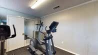 This fitness center is a dedicated, single-level exercise space designed to provide a functional and efficient workout environment. The room features a long, rectangular layout with neutral gray walls, crown molding, and a textured ceiling fitted with fluorescent lighting that ensures bright, even illumination throughout. Durable dark rubber flooring covers the entire space, offering both safety and comfort during exercise.

The facility is equipped with a selection of cardio machines, including two treadmills and an elliptical trainer, providing users with versatile options for aerobic workouts. A cable machine is also available for strength and resistance training. Large wall-mounted mirrors extend across a significant portion of the room, visually expanding the space and allowing users to monitor their form during exercise. The mirrors also reflect natural light from a window near the entrance, contributing to a brighter atmosphere.

The space is accessed through a solid entry door and includes a fire extinguisher mounted near the entrance for safety. HVAC vents are positioned throughout to maintain a comfortable climate. The overall layout is straightforward and organized, maximizing usable floor space for movement and exercise. This fitness center offers a practical and well-maintained environment for residents or guests looking to maintain an active lifestyle.