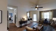 Located at 4625 Millbrook Green Dr in Raleigh, NC, this single-floor residence offers a welcoming and well-appointed living environment. The open-concept layout connects the main living area to a dining and kitchen space, creating a seamless flow throughout the home. Rich hardwood-style flooring extends across the main living area, adding warmth and a polished aesthetic to the space. A ceiling fan with integrated lighting anchors the living room, providing both comfort and illumination. Large windows dressed with curtains allow generous natural light to fill the interior, enhancing the bright and airy ambiance. A glass-panel exterior door opens to the outside, further connecting the interior to the outdoors. A counter-height bar area with seating provides a casual dining option, while a console table along one wall adds a decorative touch. The neutral wall tones and clean architectural lines give the space a timeless and versatile character. Corridor access leads to additional rooms, including what appears to be a bedroom, completing this thoughtfully designed single-level home.