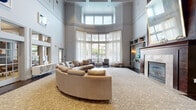 Located at 7903 Orion Circle in Laurel, Maryland, this elegantly designed communal space makes a striking first impression with its dramatic double-height ceilings and airy, open layout. The main lounge area is anchored by a generously sized curved sectional sofa set atop a patterned area rug, creating an inviting focal point beneath the soaring ceiling. A curved bay window wall floods the space with abundant natural light, filtered softly through sheer white drapery that extends from floor to ceiling, enhancing the sense of openness and volume.

Rich dark hardwood flooring runs throughout, providing a warm contrast to the cool gray and white color palette of the walls and trim. Detailed crown molding and paneled wall architecture add a refined, classical character to the contemporary interior. Built-in shelving units and display niches are thoughtfully integrated into the walls, accented by wall-mounted sconces and curated artwork, including bold abstract pieces that add visual interest.

Adjacent seating areas feature accent chairs and smaller furnishings, offering flexible arrangements for both individual use and group gatherings. French-style interior windows and glass-paned doors connect to additional rooms visible beyond, including what appears to be a dining or conference area. A fireplace with built-in cabinetry anchors one end of the room, completing this polished and welcoming community space.
