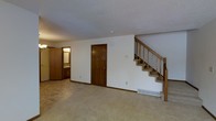 This two-floor residential space offers a practical and functional layout across both levels. The main living area is finished with wall-to-wall carpeting in neutral tones, creating a warm and comfortable atmosphere throughout. White walls are complemented by rich, dark wood trim and baseboards, adding a classic contrast that defines the interior aesthetic.

A prominent staircase with wooden railings and balusters provides access to the upper floor, serving as a central architectural feature of the main level. The staircase is finished with carpeted treads, consistent with the flooring found throughout the space.

The main level also features a closet with a dark wood-paneled door, offering practical storage. A baseboard heating unit is visible along the wall, indicating a forced-air or hydronic heating system. Adjacent to the main living area, a bathroom is visible, fitted with a vanity and mirror, providing convenient access from the common areas.

The ceilings feature a textured finish throughout, and a smoke or carbon monoxide detector is mounted on the ceiling of the main living area. The overall design of the space is straightforward and functional, with durable finishes and a neutral palette that provides a solid foundation for personalization.