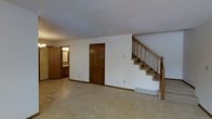 This two-floor residential space offers a practical and functional layout across both levels. The main living area is finished with wall-to-wall carpeting in neutral tones, creating a warm and comfortable atmosphere throughout. White walls are complemented by rich, dark wood trim and baseboards, adding a classic contrast that defines the interior aesthetic.

A prominent staircase with wooden railings and balusters provides access to the upper floor, serving as a central architectural feature of the main level. The staircase is finished with carpeted treads, consistent with the flooring found throughout the space.

The main level also features a closet with a dark wood-paneled door, offering practical storage. A baseboard heating unit is visible along the wall, indicating a forced-air or hydronic heating system. Adjacent to the main living area, a bathroom is visible, fitted with a vanity and mirror, providing convenient access from the common areas.

The ceilings feature a textured finish throughout, and a smoke or carbon monoxide detector is mounted on the ceiling of the main living area. The overall design of the space is straightforward and functional, with durable finishes and a neutral palette that provides a solid foundation for personalization.