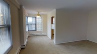 This single-floor space features a modestly sized living area finished with wall-to-wall beige carpeting that lends a warm, neutral foundation to the room. A ceiling fan with a built-in light fixture is centrally mounted, providing both air circulation and ambient lighting. Along the base of the walls, baseboard heating units run the perimeter, offering an efficient and unobtrusive heating solution.

Natural light enters the living area through two distinct window types: a standard window fitted with horizontal blinds on one wall, and a decorative glass block window that diffuses soft, natural light while maintaining privacy. The combination creates a bright yet understated atmosphere.

Crown molding accents the upper edges of the walls, adding a subtle architectural detail that elevates the overall finish of the space. The walls are painted in a clean, off-white tone throughout, contributing to an open and airy feel.

An open doorway connects the living area to the kitchen, which is partially visible and features cabinetry and what appears to be standard appliances. The layout suggests a functional flow between the main living space and the kitchen area.