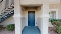 This single-story residence welcomes guests through a covered entryway framed by stucco columns and a bold teal front door, lending a sense of architectural character from the moment of arrival. The exterior reflects a Southwestern aesthetic, with textured stucco walls and desert-friendly landscaping featuring gravel groundcover, low shrubs, and towering palm trees visible from the private patio. The outdoor space offers a relaxed view of the surrounding grounds, including a gated pool area beyond the wrought-iron fencing.

Inside, the living area is thoughtfully arranged with plush carpeting underfoot and a ceiling fan overhead, contributing to a comfortable and casual atmosphere. Large windows dressed with vertical blinds allow natural light to fill the room, creating a bright and airy feel. The open layout connects the living space seamlessly to the dining area, where a pendant light fixture adds a warm focal point above the dining table. Just beyond, a partial view of the kitchen reveals light cabinetry and a raised counter, suggesting a practical and accessible layout. The overall ambiance is inviting, blending functional design with a warm, approachable character suited to everyday living.