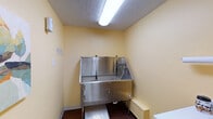 Located in Lexington, KY, this single-floor pet grooming facility — known as the Gatehouse — is thoughtfully designed to provide a clean, functional, and welcoming environment for both pets and their owners. The space features warm yellow walls throughout, complemented by rich hardwood-style flooring that lends a cohesive and polished look across all areas.

The centerpiece of the facility is a dedicated grooming room equipped with a professional-grade stainless steel grooming tub, complete with built-in restraint hooks, a handheld spray nozzle, and a non-slip diamond-plate step platform for easy pet access. Adjacent to the tub is a wooden cabinetry unit with a white countertop that provides ample workspace, along with a wall-mounted pet dryer for post-bath finishing. A paper towel dispenser and grooming supplies are conveniently within reach.

A hallway connects the grooming area to the entry, featuring a louvered interior door, a window with views to the exterior brick surroundings, and a skylight that introduces natural light into the space. Decorative artwork, including a colorful landscape painting, adds a warm and inviting touch to the corridor walls. The overall layout is compact yet highly efficient, with every element oriented toward professional pet care in a clean, comfortable setting.