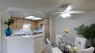 This single-story space presents an open-concept layout that seamlessly connects the kitchen and dining area, creating a cohesive and functional living environment. The kitchen is outfitted with warm wood-toned cabinetry that extends to the upper and lower sections of the walls, providing generous storage throughout. A built-in range with overhead ventilation hood anchors the cooking area, while recessed skylight panels in the kitchen ceiling introduce soft, diffused natural light. The countertops are light in finish, and a curved breakfast bar serves as a natural dividing element between the kitchen and the adjacent dining space, offering additional seating and surface area. The dining area accommodates a round glass-top table paired with white upholstered chairs, enhancing the bright and airy atmosphere. A white ceiling fan with a multi-bulb light fixture is centrally positioned overhead, providing both airflow and ambient lighting. The textured ceilings and neutral wall tones lend a classic, understated character throughout. Carpeted flooring in the dining area transitions to a smoother surface in the kitchen, and the overall layout promotes an easy flow between daily living and entertaining areas.