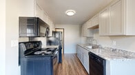 This single-floor unit offers a well-organized layout with distinct functional areas connected by light wood-look flooring throughout the main living spaces. The galley-style kitchen is a standout feature, equipped with rich dark espresso cabinetry extending to the ceiling for ample storage, marble-look countertops, a deep single-basin sink with a modern gold-toned faucet, and white appliances including a range, refrigerator, and dishwasher. A ceiling fan overhead adds airflow and light to the kitchen area, which flows naturally toward the living space.

The living area is bright and open, benefiting from multiple windows that frame views of surrounding greenery. Baseboard heating runs along the walls, and light wood-look flooring continues throughout. Bedrooms feature soft blue-grey carpet, vertical panel wall detailing, and windows that invite natural light and outdoor views. A baseboard heater in each bedroom provides individual climate control.

The bathroom is cleanly finished with dark cabinetry, a single vanity with a light countertop, a wood-framed mirror, contemporary lighting, and light wood-look flooring, offering a cohesive and updated aesthetic throughout the unit.