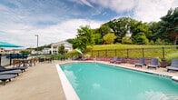 Located at 900 Reserve Way in Harrisburg, Pennsylvania, this single-story community amenity facility offers a generous footprint designed to serve residents with both fitness and recreation needs. The building is anchored by a bright, well-appointed fitness center that benefits from abundant natural light streaming through multiple large windows. The gym is equipped with cardio machines including treadmills, ellipticals, and stationary bikes, as well as a free weight rack, a weight bench, resistance mats, and a stability ball — providing a comprehensive workout environment. Motivational wall decor and a community board add a welcoming, communal touch, while recessed lighting and neutral-toned walls keep the space feeling open and clean. A tiled hallway connects the interior areas and offers a direct view through a large interior window out to the outdoor pool, creating a seamless visual connection between the indoor and outdoor amenities. The outdoor pool area is a standout feature, showcasing a large, sparkling pool surrounded by a wide concrete deck furnished with lounge chairs, seating tables, and umbrella shade options. Enclosed by a black iron fence, the pool area is framed by lush, mature trees and rolling green hillside, providing a serene, private atmosphere ideal for relaxation.