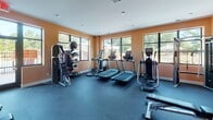 This fitness center at 139 Alexander Bank Dr. in Mooresville, NC offers a well-equipped, single-floor workout facility designed to support a full range of exercise routines. The space is characterized by warm, earth-toned orange walls that create an energizing atmosphere, complemented by a white ceiling with recessed lighting for consistent illumination throughout.

Large, multi-pane windows line the walls, flooding the interior with abundant natural light and providing views of the surrounding outdoor area, including what appears to be a community pool and landscaped grounds. Durable rubber flooring covers the entire space, offering both safety and functionality for various workouts.

The cardio zone features multiple treadmills, elliptical machines, and a stationary cycle, while the strength-training area includes a cable machine system, a weight bench, a dumbbell rack with a full set of free weights, and additional selectorized equipment. Kettlebells and a medicine ball rack add versatility for functional fitness training. A wall-mounted flat-screen television enhances the exercise experience.

Additional amenities include a water fountain station for convenience and a separate door providing easy access. The open layout ensures ample space between equipment, allowing for comfortable and unobstructed movement throughout the facility.