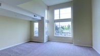 This two-floor space offers an impressive sense of openness, defined by its soaring double-height ceilings that draw the eye upward and amplify the feeling of volume. The main living area is a standout, bathed in natural light streaming through oversized windows that frame pleasant outdoor views of surrounding greenery. The generous window placement ensures the space remains bright throughout the day while maintaining a calm, airy ambiance. Soft wall-to-wall carpeting in a neutral tone provides a comfortable underfoot feel and complements the warm, light-colored walls that run throughout the interior. Subtle architectural details, including recessed ceiling elements and clean-lined beam accents, add depth and visual interest without overwhelming the space. The open layout lends itself well to a variety of furniture arrangements, offering flexibility for different uses. Ventilation is well integrated into the ceiling design, contributing to a clean and uncluttered aesthetic. The overall atmosphere is serene and inviting, with the combination of high ceilings, abundant natural light, and neutral finishes creating a versatile backdrop that is both functional and visually appealing across both levels of the space.
