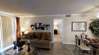 This single-story residence centers around a comfortable living area defined by wood-look flooring and a neutral, warm color palette across its walls and furnishings. The living room is anchored by a plush sofa accented with decorative pillows, complemented by a glass-top coffee table and coordinating accent pieces that add an element of elegance to the space. Crown molding runs along the ceiling perimeter, providing a refined architectural detail, while a textured popcorn ceiling adds depth overhead. Wall-mounted decorative sconces flank a framed canvas artwork, contributing to the room's carefully curated aesthetic. A large ornate mirror on the adjacent wall enhances the sense of openness. Ceiling-mounted HVAC vents ensure efficient climate control throughout. A doorway from the living area leads directly into a carpeted room, visible in the background and fitted with its own lighting and furniture, suggesting a bedroom or secondary living space. The overall ambiance is warm and inviting, with a classic, traditionally styled interior that balances functionality with decorative detail throughout the home.