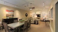 This single-floor space showcases an open-concept layout that seamlessly connects the dining and living areas, creating a cohesive and functional environment. The vaulted ceiling adds a sense of spaciousness and visual interest, while a ceiling fan with an integrated light fixture serves as a practical focal point overhead. Track lighting runs along the ceiling in multiple directions, providing flexible and well-distributed illumination throughout the space.

The dining area features a substantial rectangular table paired with upholstered chairs in a rich gray-brown finish, accommodating generous seating for gatherings. A decorative tray with greenery adds a touch of warmth to the table setting. An ornate dark-framed mirror adorns the wall nearby, contributing an elegant accent.

The adjoining living area is anchored by a neutral sofa arrangement, complemented by a dark wood coffee table and floral-patterned accent chairs. A built-in media niche houses a flat-screen television flanked by framed artwork, creating a defined focal wall. The carpeted flooring in warm beige tones unifies the space, while table lamps add soft, ambient lighting throughout.