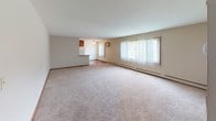 This single-floor residence offers a practical and comfortable layout across several well-defined living spaces. The expansive living room serves as the heart of the unit, featuring plush neutral carpeting, a large window with vertical blinds that fills the room with natural light, and a baseboard heating system. The open sightline to the kitchen makes the space feel connected and airy.

The kitchen is efficiently designed with warm-toned wood cabinetry, granite-look countertops, hardwood-style flooring, and white appliances including a refrigerator and range. Ample upper cabinet storage adds to its functionality.

Two bedrooms are carpeted and each equipped with a ceiling fan and window with vertical blinds, allowing for natural light and ventilation. One bedroom features a soft sage-green accent wall, adding a touch of color and warmth, while both rooms include baseboard heaters for year-round comfort.

The full bathroom is appointed with classic white subway tile surrounding the tub-shower combo, a mosaic tile floor, a white vanity with countertop, and rich dark wood cabinetry. A wood-framed entry door completes the space with a warm, traditional aesthetic throughout.