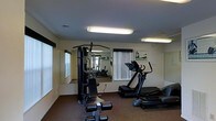 This single-floor fitness room is designed to support a well-rounded workout routine in a functional and practical setting. The space is outfitted with a multi-station cable weight machine, a treadmill, and additional cardio equipment, providing users with options for both strength training and cardiovascular exercise. A set of free weights is also available, adding further versatility to the workout options on offer.

The room features wall-to-wall mirroring along one side, which visually expands the space and allows users to monitor their form during exercise. Overhead fluorescent lighting ensures the room is brightly and evenly lit throughout. The flooring consists of a dark, durable carpet suited for fitness use, providing some cushioning underfoot.

Windows with vertical blinds line the walls, allowing for natural light to filter in while maintaining privacy. The walls are finished in a neutral off-white tone, contributing to a clean and focused environment. Framed artwork on the walls adds a modest decorative touch. The overall layout is efficient, making good use of the available floor space to accommodate multiple users and a diverse selection of equipment simultaneously.