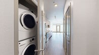 This thoughtfully designed 1-bedroom, 1-bathroom unit offers a well-appointed single-floor layout with modern finishes throughout. The open-plan living and kitchen area is anchored by a sleek galley-style kitchen featuring light wood cabinetry, stainless steel appliances including a French door refrigerator, electric range, built-in microwave, and dishwasher, complemented by a white subway tile backsplash and clean quartz countertops. Rich hardwood-style flooring flows throughout the entire unit, adding warmth to the contemporary aesthetic. A hallway off the kitchen provides access to all rooms and reveals a conveniently integrated stacked washer and dryer unit. The bedroom is a quiet, well-proportioned retreat accessed via a stylish sliding barn door. The full bathroom features large-format neutral tile surrounding a soaking tub and shower combination, a wood-toned vanity with a white countertop, and coordinating flooring. A dedicated closet provides ample storage. Large windows at the end of the main living area allow natural light to fill the space, enhancing the bright, clean atmosphere. The unit's cohesive design palette and modern fixtures make it a polished and functional living space.