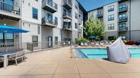 Located at 805 Canal St in Chattanooga, Tennessee, this outdoor amenity space serves as the central courtyard of a modern multi-story residential community. The focal point is a generously sized, tiled swimming pool with crystal-clear blue water, complemented by an adjacent spa. The pool deck is finished in smooth concrete and outfitted with multiple cushioned lounge chairs and umbrellas, providing ample space for sunbathing and relaxation. A pergola-style shaded seating area offers a comfortable retreat from the sun, while raised planter beds featuring vibrant flowering plants and greenery add natural color and texture throughout the space. Decorative Cor-Ten steel planters overflow with trailing foliage, contributing to the courtyard's contemporary aesthetic. The surrounding multi-story building with balconies frames the space on multiple sides, creating a private, sheltered atmosphere. Mature trees and carefully maintained landscaping, including hostas, evergreen shrubs, and seasonal blooms, line the perimeter walkways, enhancing the sense of a lush urban oasis. The overall ambiance is bright, well-maintained, and inviting, designed to offer residents a resort-like outdoor experience in the heart of Chattanooga.