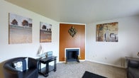 This single-floor space centers around a comfortable living area that blends retro-inspired design with a clean, neutral aesthetic. The focal point of the room is a wood-paneled accent wall finished in warm amber tones, which frames a classic fireplace with a white surround and dark firebox. A diamond-shaped metallic wall sculpture in gold and black is mounted above the fireplace, adding a decorative mid-century modern flair to the feature wall.

The surrounding walls are painted in a crisp white, providing a bright contrast to the warm tones of the accent wall. Soft, wall-to-wall carpeting in a light beige runs throughout the living area, contributing to the room's cozy atmosphere. A textured popcorn ceiling adds a characteristic period detail to the space.

Artwork adorns the walls on either side of the fireplace, complementing the room's casual yet curated style. A sleek black side table positioned near a leather sofa offers functional surface space. The overall layout feels open and well-proportioned, with thoughtful design elements that create a warm and welcoming living environment suited for relaxed, everyday living.