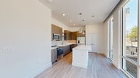 Located at 2351 Plaza Blvd in Richardson, Texas, this contemporary two-story residence offers three bedrooms and three bathrooms across a well-planned layout. The main floor hosts the primary living areas, featuring an open-concept kitchen and living room flooded with natural light through floor-to-ceiling glass doors that lead to an outdoor balcony. The kitchen is equipped with stainless steel appliances, including a dishwasher, microwave, and refrigerator, complemented by wood-toned cabinetry, a subway tile backsplash, white quartz countertops, and a central island with pendant lighting overhead. Light hardwood-style flooring runs throughout the main level, contributing to the bright, airy atmosphere. A ceiling fan accents the living area, which benefits from abundant views of surrounding greenery. The bathrooms showcase clean, modern finishes, including a glass-enclosed walk-in shower tiled in marble-style white stone with brushed nickel hardware, and additional bathrooms featuring wood-look flooring with gold-toned fixtures. The upper floor contains the bedrooms, including a primary suite with plush carpeting, a tray ceiling, an en-suite bathroom, and a dedicated walk-in closet. Additional bedrooms also feature soft carpeting and recessed lighting, providing comfortable and versatile spaces throughout.