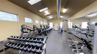 This fitness center offers a comprehensive workout environment designed to accommodate a wide range of exercise needs. The space is bright and airy, benefiting from a row of skylights along the ceiling that flood the room with natural light, complemented by overhead fluorescent fixtures for consistent illumination throughout all hours of operation.

The layout is thoughtfully organized, with a dedicated cardio zone featuring a lineup of professional-grade treadmills, ellipticals, and upright bikes—predominantly Cybex-brand equipment—arranged in a row along one side of the room. A large wall-mounted mirror enhances the sense of space and allows users to monitor their form during workouts.

The free weights area is well-stocked with a full dumbbell rack offering a wide range of weights, neatly positioned for easy access. Additional strength and resistance training equipment rounds out the offerings, providing options for full-body workouts.

The flooring features durable rubber gym matting, ideal for heavy equipment and high-impact activity. Warm beige walls and a vaulted ceiling with an exposed ventilation duct give the space an industrial yet welcoming character. Small windows along the perimeter allow additional natural light to enter. A ceiling fan provides supplemental air circulation, contributing to a comfortable training environment overall.