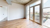 Located at 2200 Market St in Denver's vibrant Five Points neighborhood, this two-bedroom, one-bathroom loft-style residence showcases an industrial-chic aesthetic defined by exposed concrete ceilings, visible ductwork, and raw concrete columns throughout. The open-concept kitchen and living area serves as the heart of the home, featuring warm hardwood flooring, stainless steel appliances, white subway tile backsplash, sleek white cabinetry, and a large waterfall-edge island with an undermount sink and matte black fixtures illuminated by pendant lighting. The living area opens to a private balcony through full-height French doors, flooding the space with natural light and offering elevated views of the Denver skyline. The primary bedroom is generously proportioned with plush carpeting, a large window with city views, and an exposed concrete accent column. The second bedroom also features hardwood floors, a built-in closet, and exposed architectural ceiling elements. The full bathroom is finished with warm wood-toned cabinetry, a double-sink vanity with matte black fixtures, a large mirror, and a soaking tub with shower. Track lighting and an industrial palette tie the entire residence together into a cohesive, modern urban living experience.