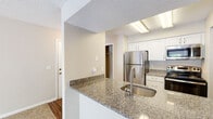 Located at 103 Melville Loop in Chapel Hill, NC, this single-floor residence offers a well-appointed layout with a clean, neutral aesthetic throughout. The kitchen is a functional highlight, featuring granite countertops, white cabinetry, a stainless steel refrigerator, electric range, over-the-range microwave, and a breakfast bar with an undermount sink — ideal for both cooking and casual dining. The living area is carpeted and generously sized, anchored by a classic wood-mantel fireplace with a dark tile surround. A sliding glass door opens to a private balcony with a white railing, providing a view of the surrounding greenery. The bedroom is comfortably proportioned with plush carpet, natural light from a window, and access to a closet. The bathroom includes a tiled tub-shower combo, granite vanity, and wood-look flooring. A dedicated laundry area houses a full-sized top-loading washer with overhead shelving for added storage convenience. Textured ceilings and warm neutral wall tones create a consistent, cohesive atmosphere throughout the space, making it a comfortable and practical home in a wooded Chapel Hill setting.