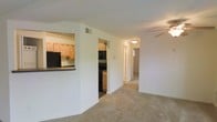This single-floor residence is designed with an open and functional layout that connects the main living areas seamlessly. The living area is finished with wall-to-wall carpeting in a neutral tone, creating a warm and comfortable atmosphere. A ceiling fan is mounted overhead, adding both practical ventilation and a subtle decorative element to the space.

The kitchen is positioned adjacent to the living area and is accessible via a pass-through opening, which enhances the sense of openness between the two spaces. It features light wood cabinetry offering ample storage, dark countertops, and black appliances including a microwave and range, providing a clean contrast against the lighter finishes. The kitchen also has a separate entry point, allowing for convenient access from multiple directions.

A hallway extends from the main living space, leading toward additional rooms at the rear of the residence. The corridor is well-lit with a flush ceiling light, and the view down the hall suggests further living quarters beyond. Throughout the space, the walls are painted in a clean, neutral white, contributing to a bright and airy feel. The overall design is straightforward and practical, making efficient use of the available floor plan.