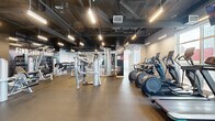 The Line Fitness Centre, located in Surrey, BC, is a professionally designed facility offering a comprehensive workout environment across a single, expansive floor. The space features an industrial-chic aesthetic, with exposed ductwork and ventilation systems running across dark ceilings, offset by bright linear pendant lighting that illuminates the floor below.

The main training area is generously proportioned, accommodating a wide array of cardio equipment including treadmills, ellipticals, and stair climbers, all positioned along large floor-to-ceiling windows that allow natural light to filter in and offer outdoor views. A dedicated strength and free weights zone features cable machines, a squat rack, kettlebell storage, adjustable benches, and a full dumbbell rack, all set on durable rubber flooring. Motivational graphics and bold typographic wall art reinforce the energetic atmosphere throughout.

A separate functional training room is enclosed with glass partitions, providing a semi-private space for focused workouts. A hydration station is conveniently located near the entrance, alongside glass double doors for easy access. The facility also includes a clean, accessible bathroom finished with light tile flooring, a wall-mounted sink, and grab bar supports. The overall design balances functionality with a modern, high-energy aesthetic suited to a dedicated fitness environment.
