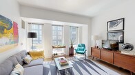 This single-floor residence offers a thoughtfully designed layout that balances contemporary style with everyday functionality. The living area is a standout space, characterized by an abundance of natural light streaming through a bank of large, gridded windows that frame urban views of the surrounding cityscape. White walls and light hardwood flooring create a clean, airy backdrop, while the open layout allows for flexible furnishing arrangements. A bold abstract artwork anchors one wall, adding a vibrant focal point to the room.

The kitchen is efficiently designed with soft grey cabinetry accented by brass hardware, white quartz countertops, and a striking marble-patterned tile backsplash. A central island with bar seating provides additional prep space and casual dining. Full-sized white appliances and generous cabinet storage complete the practical layout, illuminated by a sleek brass pendant fixture overhead.

The bedroom is quietly tucked away, benefiting from large windows that invite natural light and offer outdoor views. A double-door closet provides ample storage. The full bathroom features dark subway tile surrounding a full soaking tub, dark floor tile, and a stacked in-unit washer and dryer conveniently integrated within the space.
