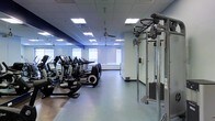 This fitness center offers a thoughtfully designed workout environment spread across a single floor. The space is illuminated by a grid of recessed panel lighting set within a drop-tile ceiling, complemented by natural light streaming through large windows along the far wall, creating a bright and energizing atmosphere. Ceiling fans are installed throughout, providing additional air circulation during intense workouts.

The cardio zone features a row of Life Fitness stationary bikes and elliptical machines, all equipped with individual digital screens for performance tracking. These are positioned along one side of the floor, which is finished with durable, cushioned fitness flooring ideal for high-traffic use. Adjacent to the cardio equipment stands a multi-station cable and resistance training machine, suitable for a wide variety of strength exercises.

Full-length mirrors line one wall, enhancing the sense of openness and allowing users to monitor their form during exercise. The overall color palette — composed of cool blues, whites, and grays — contributes to a clean, modern aesthetic. The layout is open and spacious, allowing for comfortable movement between equipment stations. This fitness center is well-suited for both cardio and strength training routines in a professional, facility-grade setting.