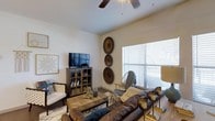 This single-floor space is thoughtfully designed to offer comfort and style in equal measure. The living area serves as the heart of the space, showcasing a warm, earthy aesthetic with a rich leather sofa as the centerpiece, complemented by a woven rattan coffee table and layered area rug that add natural texture. A media cabinet with glass-front doors houses the flat-screen television, while a curated arrangement of decorative circular wall art adds a distinctive, bohemian-inspired focal point along the neutral-toned walls. A wicker accent chair and additional seating provide flexible arrangements for gathering or relaxing. Natural light flows generously into the room through a large window dressed with horizontal blinds, brightening the light-colored walls and creating an airy, open feel. A ceiling fan overhead ensures year-round comfort while contributing to the room's casual, relaxed character. The smooth white ceiling and clean architectural lines give the space a timeless, uncluttered foundation that allows the warm, layered furnishings and décor to take center stage. Overall, the space presents a cohesive and inviting environment that balances practical functionality with a distinctive, curated sense of style.