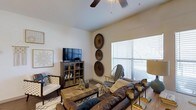 This single-floor space is thoughtfully designed to offer comfort and style in equal measure. The living area serves as the heart of the space, showcasing a warm, earthy aesthetic with a rich leather sofa as the centerpiece, complemented by a woven rattan coffee table and layered area rug that add natural texture. A media cabinet with glass-front doors houses the flat-screen television, while a curated arrangement of decorative circular wall art adds a distinctive, bohemian-inspired focal point along the neutral-toned walls. A wicker accent chair and additional seating provide flexible arrangements for gathering or relaxing. Natural light flows generously into the room through a large window dressed with horizontal blinds, brightening the light-colored walls and creating an airy, open feel. A ceiling fan overhead ensures year-round comfort while contributing to the room's casual, relaxed character. The smooth white ceiling and clean architectural lines give the space a timeless, uncluttered foundation that allows the warm, layered furnishings and décor to take center stage. Overall, the space presents a cohesive and inviting environment that balances practical functionality with a distinctive, curated sense of style.