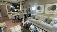 This single-floor residence showcases a thoughtfully curated open-concept layout where the living, dining, and kitchen areas flow seamlessly into one another. The living area is anchored by a plush, light gray sofa adorned with an array of decorative pillows in sage green, taupe, and soft white tones, complemented by a rounded dark wood coffee table styled with candles, fresh florals, and accent décor. A geometric patterned area rug grounds the seating area, while a large floor mirror and statement wall art featuring botanical motifs add depth and visual interest to the space. The dining area sits adjacent to the living room and features a sleek dark wood table surrounded by upholstered gray chairs, creating an elegant yet inviting setting for entertaining. Organic decorative elements, including sculptural moss balls and twisted branch arrangements, are woven throughout the space, bringing a natural, earthy quality to the interior. The kitchen, visible in the background, features dark cabinetry, pendant lighting, and a mosaic tile backsplash, offering a rich contrast to the warmer tones of the living and dining areas. Together, the spaces are unified by a warm taupe and sage green palette, track lighting, and carefully chosen furnishings that create a polished, welcoming ambiance.