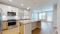 Located at 1650 Clearwater Largo Rd in Largo, Florida, this single-floor residence showcases a clean, contemporary design with thoughtful finishes throughout. The open-concept kitchen is a standout feature, offering granite countertop surfaces, a central island with pendant globe lighting, and a full suite of stainless steel appliances including a refrigerator with ice maker, range, built-in microwave, and dishwasher. Cabinetry combines crisp white upper cabinets with warm natural wood lower cabinets, accented by matte black hardware, while a white subway tile backsplash adds a polished finishing touch. Light hardwood-style flooring runs throughout the main living areas, seamlessly connecting the kitchen to an open living space that benefits from natural light through large windows and a ceiling fan overhead. A hallway leads to the bedroom, which features soft carpet, neutral walls, and a window that invites additional natural light. The bathroom is well-appointed with a combined tub and shower, a wood-toned vanity with granite countertop, and clean white tile surround. The overall aesthetic throughout the space is bright, modern, and cohesive, with a neutral color palette that creates a calm and welcoming atmosphere.