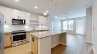 Located at 1650 Clearwater Largo Rd in Largo, Florida, this single-floor residence showcases a clean, contemporary design with thoughtful finishes throughout. The open-concept kitchen is a standout feature, offering granite countertop surfaces, a central island with pendant globe lighting, and a full suite of stainless steel appliances including a refrigerator with ice maker, range, built-in microwave, and dishwasher. Cabinetry combines crisp white upper cabinets with warm natural wood lower cabinets, accented by matte black hardware, while a white subway tile backsplash adds a polished finishing touch. Light hardwood-style flooring runs throughout the main living areas, seamlessly connecting the kitchen to an open living space that benefits from natural light through large windows and a ceiling fan overhead. A hallway leads to the bedroom, which features soft carpet, neutral walls, and a window that invites additional natural light. The bathroom is well-appointed with a combined tub and shower, a wood-toned vanity with granite countertop, and clean white tile surround. The overall aesthetic throughout the space is bright, modern, and cohesive, with a neutral color palette that creates a calm and welcoming atmosphere.