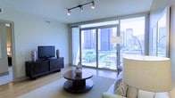 This contemporary single-floor residence is thoughtfully designed to maximize both style and comfort in an urban setting. The living area serves as the centerpiece of the space, anchored by expansive floor-to-ceiling sliding glass windows and doors that flood the interior with natural light and offer breathtaking, unobstructed views of a dynamic city skyline. The seamless connection to an outdoor balcony further extends the living space, inviting the energy of the city indoors.

The living room is finished with light hardwood flooring that adds warmth and continuity throughout. A sleek, dark wood media console provides a grounded contrast to the bright, airy surroundings, while a round wooden coffee table and neutral area rug create an inviting, well-composed seating arrangement. Track lighting on the ceiling provides flexible, directional illumination to complement the abundant natural light.

The overall ambiance is modern and sophisticated, blending clean architectural lines with carefully chosen furnishings. The interplay of natural light, city views, and refined interior details gives this space a cosmopolitan character that is both functional and visually compelling — ideal for those who appreciate contemporary urban living.