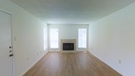 This single-floor space offers a straightforward and functional layout with a focus on clean design and practical finishes. The main living area serves as the centerpiece of the space, featuring warm hardwood-style flooring that extends throughout the room, providing a cohesive and easy-to-maintain surface underfoot.

A tile-surround fireplace anchors one wall of the living area, offering both a functional heating element and a subtle architectural focal point. The neutral tile surround complements the light-toned walls, contributing to the room's understated aesthetic.

Two windows flank the fireplace symmetrically, allowing natural light to fill the space and creating a bright, airy atmosphere. The white walls and light-colored trim throughout reinforce the open, neutral feel of the room.

The ceiling features a textured finish and sits at a standard height, framing the room in a comfortable and proportionate manner. White baseboards line the perimeter, adding a clean, defined boundary between the walls and flooring.

Overall, the space presents a blank canvas well-suited for a variety of furnishing styles, with its neutral palette and practical finishes making it a versatile and move-in-ready environment.