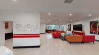 Avoa Luxury Living presents a striking, contemporary communal space defined by bold design choices and a vibrant color palette. The entry lobby makes an immediate impression with its crisp white walls, gleaming large-format tile flooring, and the prominently displayed Avoa Luxury Living logo that anchors the reception area. Signature red accent stripes and a curated arrangement of bold red and orange seating create a dynamic, energetic atmosphere throughout the lounge area. A large flat-screen display and carefully selected wall art, including vivid figurative pieces, add cultural depth to the space.

Moving deeper into the property, a secondary lounge area showcases soaring double-height ceilings and floor-to-ceiling glass walls that frame views of lush exterior greenery, bathing the interior in abundant natural light. A generously sized curved charcoal leather sofa serves as the centerpiece, complemented by colorful accent pillows and geometric floor cushions in fuchsia and orange. Contemporary abstract artwork adorns the bright white walls, reinforcing the space's artistic identity. The seamless blend of natural light, modern architecture, and carefully curated furnishings creates an upscale, welcoming ambiance that reflects the luxury living experience Avoa aims to deliver.