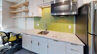 This thoughtfully designed single-floor studio unit offers a compact yet functional open-plan layout that seamlessly integrates living, dining, and sleeping areas. The kitchenette is a standout feature, showcasing eye-catching chartreuse green subway tile backsplash, sleek white cabinetry, light stone countertops, a stainless steel sink, and an overhead microwave — creating a modern and energetic focal point. The living area transitions smoothly from tile to plush carpet with vibrant chartreuse accent panels, and is furnished with a dark leather sofa and a clean white coffee table, providing a comfortable lounge space. The sleeping area is anchored by a built-in platform bed with an integrated wood headboard featuring open shelving and adjustable reading lamps. Floor-to-ceiling windows flood this area with abundant natural light and offer views of the surrounding urban environment. Track lighting throughout adds to the bright, contemporary atmosphere. The bathroom is finished with neutral grey textured tile on the walls and floor, a wall-mounted pedestal sink, a large mirror with vanity lighting, and a spacious walk-in shower with a built-in niche and handheld fixture — combining clean aesthetics with practical functionality throughout.