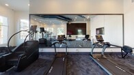 Avidity Fitness in Lutz, Florida is a well-appointed, single-story fitness facility designed to accommodate a comprehensive range of workout needs. The space is organized into distinct functional zones, each thoughtfully equipped and finished to support both focused training and group activity.

The main workout area features soaring ceilings accented by bold navy geometric ceiling details and recessed lighting, creating a bright and energizing atmosphere. Large windows along multiple walls invite generous natural light and offer views of the surrounding palm-lined exterior. Patterned blue-toned carpet tiles cover the floors throughout the primary exercise zones, lending a modern, durable finish. This area is equipped with cable machines, a multi-functional rack system, weight benches, and a full dumbbell rack, supporting a wide variety of strength training exercises.

The cardio section houses a row of True treadmills, stair climbers, and additional cardio equipment, all positioned facing large wall-mounted mirrors and flat-screen televisions. The mirrored walls visually expand the space and allow users to monitor their form during workouts.

A separate studio room provides a clean, open environment finished with light hardwood-style flooring, a ceiling fan, recessed linear lighting, full-length mirrors with a ballet barre, and large windows overlooking greenery. The entry area offers high ceilings, glass-panel doors, and open sightlines into the main facility, creating a welcoming and transparent arrival experience.