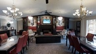 This sophisticated single-floor lounge space exudes an upscale, club-like ambiance through its carefully curated design elements. The centerpiece of the room is a built-in entertainment unit framing a flat-screen television and a sleek fireplace below, flanked by dark wood cabinetry that lends a sense of structure and elegance. On either side, rich red tufted leather banquette seating lines the walls, offering generous and plush accommodations for guests. A large black leather ottoman anchors the seating arrangement at the center of the room, complemented by an eclectic mix of accent chairs in red and neutral tones. Two ornate crystal chandeliers hang symmetrically, casting warm, ambient light throughout the space, while a ceiling fan at the apex adds a practical touch. The vaulted ceiling contributes to a sense of openness and volume. Decorative wall art and collectible display pieces add personality and visual interest to the dark, richly toned walls. The bold leopard-print carpeting ties the room together, reinforcing the lounge's daring and distinctive aesthetic. Overall, the space balances dramatic décor with comfortable furnishings, creating an inviting atmosphere well-suited for socializing and relaxation.