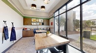 Located in Mesa, AZ, this single-floor Makers Space offers a thoughtfully designed combined kitchen and dining area built for creativity and collaboration. The room features elevated ceilings with recessed lighting and stylish drum pendant fixtures that contribute to an open, airy atmosphere. Horizontal color-blocked wall stripes in sage green, blush pink, and white add a distinctive, artistic character throughout the space.

Sturdy butcher block worktables with industrial-style metal bases and modern seating provide ample workspace for multiple users. Along one wall, warm wood cabinetry—including upper cabinets and open display shelving—frames a sleek gray countertop equipped with a professional-grade spring-neck faucet and an undermount sink, ideal for hands-on projects. The terrazzo-style flooring adds a polished, durable finish underfoot.

Floor-to-ceiling black-framed windows flood the space with natural light and offer clear views of an outdoor courtyard and playground area beyond. Wall-mounted apron hooks and tasteful sculptural wall art reinforce the functional yet creative spirit of the room. A glazed entry door provides easy access, completing a space that balances practical utility with modern, inviting design.