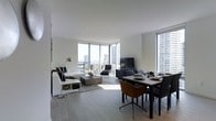This contemporary single-floor residence showcases a sleek, open-concept layout that seamlessly integrates the living and dining areas into one cohesive, light-filled space. The interior is defined by clean lines, a neutral white and grey color palette, and light hardwood-style flooring that runs throughout, lending a sense of warmth and continuity.

Floor-to-ceiling windows wrap around the living area, inviting an abundance of natural light and framing impressive panoramic views of the city skyline and beyond. The living space is anchored by a comfortable sofa ensemble, a stylish low-profile coffee table, and a flat-screen television, all arranged to take full advantage of the spectacular outlook. A chaise lounge positioned near the window further emphasizes the light and views.

Adjacent to the living area, the dining space accommodates a rectangular table with a set of modern chairs in contrasting dark tones, creating a sophisticated entertaining area. The overall ambiance is polished and minimalist, with carefully selected furnishings that complement the architectural design without overwhelming the sense of openness. The abundance of natural light, high vantage point, and thoughtful layout combine to create an elegant and functional urban living environment.
