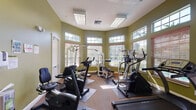This fitness center is housed within a single-level space designed to support a well-rounded workout experience. The room features a distinctive angled layout with large windows lining multiple walls, fitted with wood-toned blinds and topped with transom windows that allow abundant natural light to fill the space. The olive-green walls contribute a clean, energizing atmosphere, while the textured ceiling with recessed panel lighting ensures consistent illumination throughout.

The flooring consists of a combination of carpet and rubber exercise mats strategically placed beneath the equipment to provide traction and protection. The cardio section is well-stocked, featuring treadmills, elliptical trainers, and upright and recumbent stationary bikes. A wall-mounted flat-screen television is positioned for visibility from the cardio machines, enhancing the workout experience. The strength-training area includes a multi-station cable and weight machine, offering a range of resistance exercises.

Access to the room is provided through both a standard door and a set of French doors, adding an open, welcoming character. Informational postings are visible on the walls, providing guidelines for facility use. The overall ambiance is bright, functional, and well-maintained.