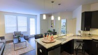 This single-floor space showcases a thoughtfully designed open-concept layout that integrates the kitchen, dining, and living areas into one cohesive environment. The kitchen serves as a focal point, anchored by a large granite-topped island with a built-in sink and undermount basin, offering ample workspace and informal seating for several guests. Dark cabinetry contrasts elegantly with the light granite countertops, while a stainless steel dishwasher adds a modern, functional touch.

Three sleek pendant lights hang above the island, providing focused task lighting and contributing to the contemporary aesthetic of the space. The open floor plan allows for a smooth transition into the dining area, which is furnished with dark chairs, and further into the living room, where hardwood-style flooring and a patterned area rug add warmth and texture.

Large windows and a glass-paneled door flood the living area with natural light, enhancing the bright, airy atmosphere throughout. A glimpse into an adjoining bedroom reveals additional living space, suggesting a well-connected and functional floor plan. The overall ambiance is modern, clean, and inviting.