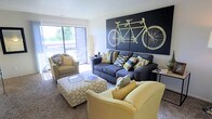 This single-story residence is thoughtfully designed with a cohesive color palette of navy, gold, and neutral tones carried throughout the space. The living room serves as the focal point of the home, anchored by a bold, large-scale black wall art panel featuring a tandem bicycle illustration, adding a striking graphic element to the room. Comfortable seating, including a navy sofa and mustard-yellow accent chairs, surrounds a patterned ottoman that doubles as a coffee table. A sliding glass door fills the living area with natural light and provides access to an outdoor balcony, offering views of surrounding greenery. Soft carpet flooring extends throughout the living and sleeping areas.

The kitchen and dining area feature tile-style flooring and include standard appliances — a white range, refrigerator, and wood-toned cabinetry. A dark dining table with upholstered chairs provides a casual yet refined dining experience.

The bedrooms are carpeted and well-lit, each with large windows that bring in ample natural daylight and outdoor views. One bedroom features a spacious closet, while both rooms offer comfortable layouts suitable for full-sized furnishings. The overall ambiance is modern, coordinated, and welcoming.
