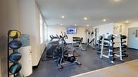 This fitness room offers a comprehensive workout environment designed to support a wide range of exercise routines. The space features recessed lighting set into a clean white ceiling, complemented by natural light filtering in through windows along one wall, creating a bright and energizing atmosphere. The floor is finished with durable rubber matting, providing both safety and functionality for various types of training. Cardio equipment is prominently positioned, including an air resistance bike and a stair climber, ideal for high-intensity interval training. A dedicated free weights area includes a multi-tiered dumbbell rack stocked with a full range of weights, accompanied by an adjustable bench for versatile strength training. A cable machine station adds the ability to perform a broad variety of resistance exercises, while a barbell and weight plate station rounds out the strength training options. A wall-mounted flat-screen television is positioned for easy viewing from multiple points in the room, adding an entertainment element during workouts. The overall layout is organized and spacious, allowing users to move freely between stations without congestion.
