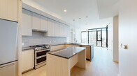 Located at 675 9th Ave in San Diego's vibrant Little Italy neighborhood, this contemporary single-floor residence showcases a thoughtfully designed open-concept layout with clean lines and modern finishes throughout. The kitchen serves as the centerpiece of the living space, featuring a large island topped with striking dark granite countertops, flat-front cabinetry in light wood and matte gray tones, and a classic subway tile backsplash. Premium stainless steel appliances include a double-oven gas range, a built-in microwave, and a full-size French door refrigerator, making this kitchen as functional as it is stylish. Pendant lighting above the island adds a refined touch. The living area flows seamlessly from the kitchen, offering light hardwood flooring, built-in open shelving, and an exposed concrete structural column that adds an industrial character. Floor-to-ceiling windows and glass doors flood the space with abundant natural light while framing views of the surrounding cityscape. The bathroom features large-format marble-look tile wrapping the walls of a deep soaking tub and shower combination, complemented by brushed gold fixtures and a built-in niche for a polished, spa-like finish. Overall, the space presents a cohesive blend of modern urban design and refined comfort.
