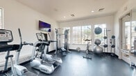 Located at 9955 Buffalo Speedway in Houston, TX, this fitness center offers a well-equipped exercise space designed to support a variety of workout routines. The room features durable dark rubber flooring throughout, recessed lighting in the ceiling, and multiple windows along the walls that allow ample natural light to fill the space while providing views of the surrounding outdoor area, including a glimpse of a pool beyond the glass door exit.

The cardio section is stocked with True-brand treadmills, elliptical cross-trainers, and a stationary cycling bike, catering to a range of aerobic exercise preferences. A wall-mounted flat-screen television is positioned for easy viewing during workouts. A multi-function cable resistance machine with an attached bench serves as the centerpiece for strength training, complemented by a rack of hex dumbbells in graduated weights and additional weight plates stored nearby. Stability and medicine balls of varying sizes add flexibility for functional training exercises.

Full-length mirrors line one wall, visually expanding the space and allowing users to monitor their form during workouts. A direct door exit leads to the outdoor area, enhancing ventilation and accessibility. The clean, white walls and efficient layout make this fitness center a practical and functional amenity for residents at this Houston location.