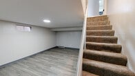 This basement level has been thoughtfully finished to provide a clean, functional living space. The main open area features luxury vinyl plank flooring in a warm grey wood-tone finish, complemented by neutral grey-painted walls and dark baseboards that create a cohesive, modern aesthetic. A circular flush-mount ceiling light provides overhead illumination, while a small window along one wall allows a modest amount of natural light to filter in. Wainscoting-style paneling adds a subtle decorative detail along the lower portion of one accent wall.

A carpeted staircase with a white-painted stringer and open railing connects the basement to the upper level, and the underside of the stair is incorporated cleanly into the layout. A separate utility room, accessible through a doorway adjacent to the main space, houses the mechanical systems including a water heater and electrical panel, keeping these functional elements contained and out of the primary living area.

An additional room off the main space mirrors the same flooring and wall finishes, featuring its own window and ceiling light fixture, making it suitable for use as a bedroom, office, or flex space. Overall, the basement presents a clean, move-in ready environment with practical utility access and versatile open areas.